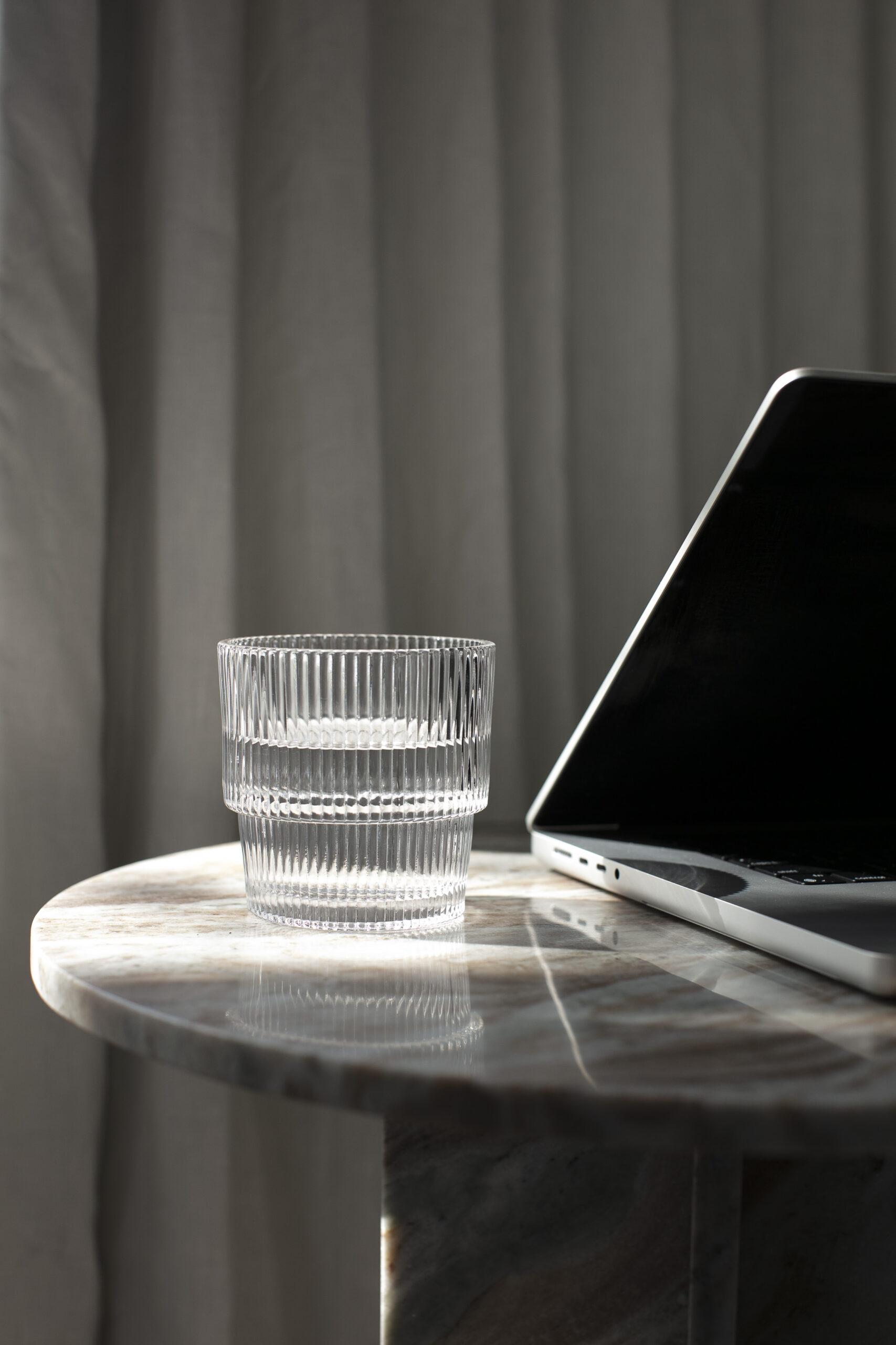 Laptop partially open and glass of water on a round marble table with a dark and moody vibe